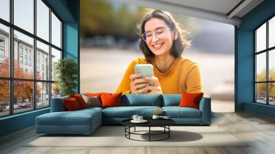 Smiling young woman sitting outside using her phone Wall mural