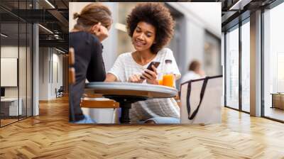 two young woman sitting at outdoor cafe together and looking at mobile phone Wall mural