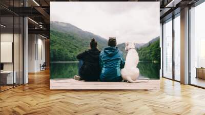 Couple sitting with a dog on dock at the lake. Back view Wall mural