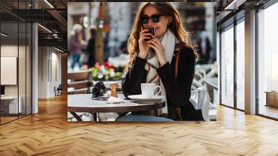 Smiling young woman is using her smartphone while enjoying a coffee break at an outdoor cafe Wall mural