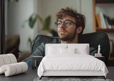 Pensive young man wearing glasses is sitting and relaxing at home Wall mural