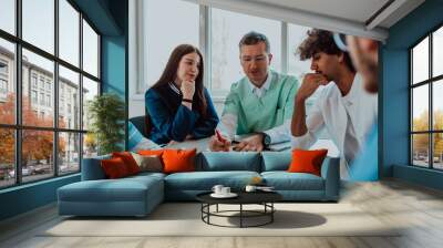 A group of multiethnic medical professionals including doctors, surgeons, and nurses are gathered in a hospital setting discussing patient care and using modern technology to address challenges in Wall mural