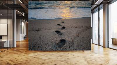 Footprints on a sandy beach disappearing into the sea at sunset. Wall mural