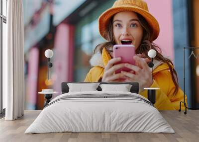 A surprised young lady using a cell phone with a positive expression, wearing a yellow jacket and hat. Happy and adorable woman, rejoicing in success Wall mural