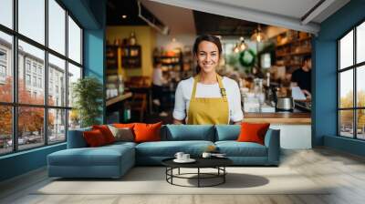 A cheerful barista wearing a yellow apron, holding a tablet in a warm and inviting cafe, ready to serve customers Wall mural