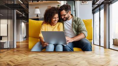 A happy couple smiling and using a laptop together while sitting on a yellow couch at home Wall mural