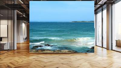 Ocean Waves Crashing Against Rocky Shore With Swimmers In The Blue Sea On A Clear Day Wall mural