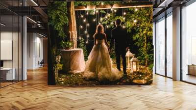 Night wedding ceremony with candles, lanterns and lamps on tree. Bride and groom holding hands on background of baulb lights, back view. Beautiful young couple standing under a tree at night Wall mural