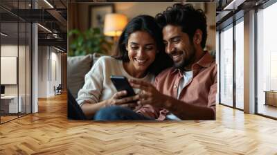 indian couple smiling and looking at phone sitting on sofa at home Wall mural