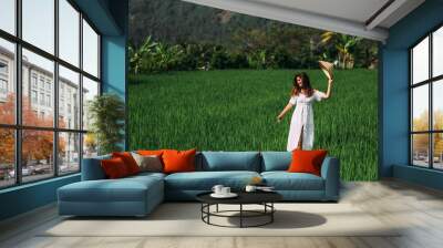 A beautiful woman walks on rice terraces on the island of Bali. A beautiful woman in a white dress and a conical hat walks through green rice fields. Tourist traveling in Indonesia. Copy space Wall mural