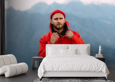 Portrait of a brutal bearded man in a red jacket and hat among the mountains. Male portrait on the background of a mountain landscape. A traveler on the background of mountains. Wall mural