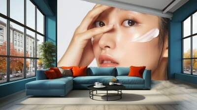 Asian woman applies facial moisturizer makeup on face in studio setting with relaxed atmosphere. Dark hair styled neatly, wearing white shirt. Focus on face with cream. Wall mural