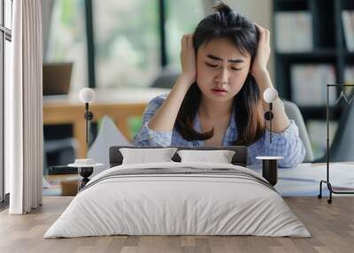 Asian young woman sits at desk, staring at financial documents in frustration. Calculating machine lies idle beside. Concerned expression on face shows stress from calculating expenses on bills, Wall mural