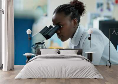 Black female scientist examines object under microscope in laboratory. Woman in white lab coat focuses on experiment with hand on eyepiece. Equipment and computer monitor in blurred background. Wall mural