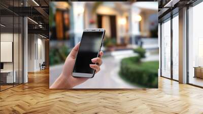 Female hand holding smartphone on blurred house background. Close-up of hand, modern home security system concept. Camera, cctv, and digital surveillance technology for home protection and safety. Wall mural