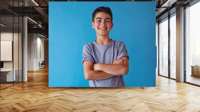 Teenage boy with happy face, smiling with crossed arms, standing over blue isolated background. Attractive, casual attire, studio portrait of young man. Wall mural