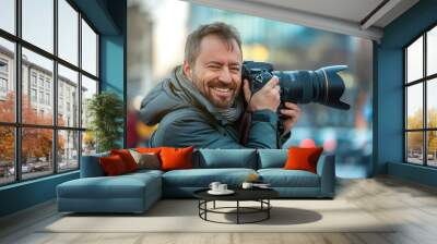 Urban photographer stands out in cityscape, smiling and holding camera high. Man in black jacket and scarf captures the world around him, with cars and buildings creating a dynamic background. Wall mural