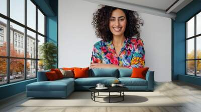 Young arab woman with curly hair, wearing a colorful floral shirt, stands confidently in a white background, smiling with crossed arms, looking directly at the camera. Wall mural