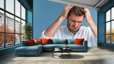 Young handsome man suffers from headache, desperate, stressed due to migraine pain. Hands on head, isolated background. Expressing tension, worry, frustration. Wall mural