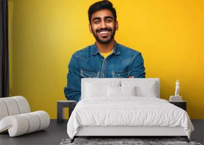 Young Indian man in casual attire, wearing denim shirt, standing on isolated yellow background. Happy face smiling with crossed arms, looking confident and cheerful at the camera. Wall mural