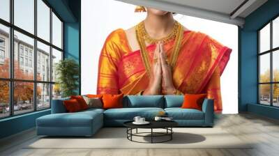 Young woman in vibrant red and gold saree stands confidently against white background. Gold jewelry, prayer position, colorful attire in focus. Simple background highlights woman beauty. Wall mural