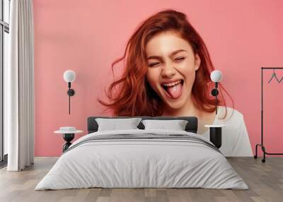 Young woman smiling with tongue out, winking at camera. Isolated on bright pink background. Happy, cheerful, caucasian girl with casual attire. Expressing joy, positive emotions. Wall mural