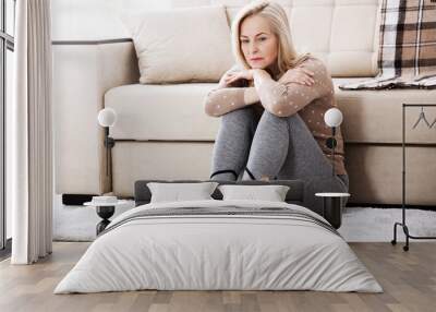 Middle aged barefoot woman sitting at the floor embracing her knees, near sofa at home, her head down, bored, troubled with domestic violence. Wall mural