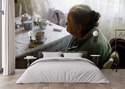 Old depressed woman. An elderly lonely woman sits at a table in the kitchen near the window and drinking tea. Wall mural