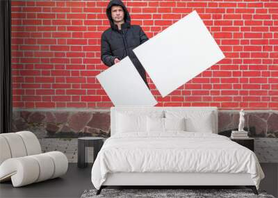 A young man stands against a background of a red brick wall and holds in front of him two white blank canvases for painting Wall mural