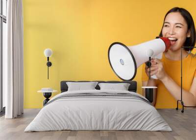 Announcement. Happy asian woman shouting loud at megaphone, recruiting, protesting with speaker in hands, standing over yellow background Wall mural