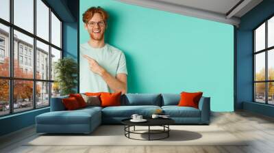 Attractive young man with red beard and hair pointing finger at blank smartphone screen, showing online promotion or app, smiling at camera, turquoise background Wall mural