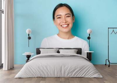 Close-up of cheerful carefree asian girl in white t-shirt smiling broadly. Pleasant korean female looking friendly at camera, enjoying nice conversation, talking with someone, light blue background Wall mural