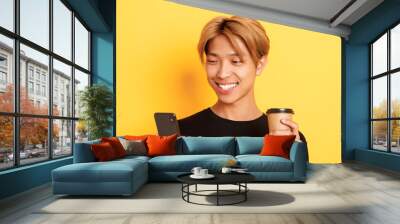 Close-up of handsome stylish asian male student, using mobile phone and drinking coffee. Guy messaging on smartphone, standing over yellow background Wall mural