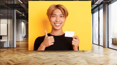 Close-up of satisfied asian guy recommend bank, showing credit card and standing over yellow background Wall mural