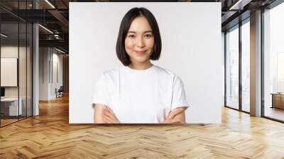 Close up portrait of confident korean girl, student looking at camera with pleased smile, arms crossed on chest, standing over white background Wall mural