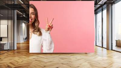 Close-up portrait of friendly joyful blond girl in white dress, sending positive vibes, smiling pleased and wink, stick tongue showing peace sign, looking kawaii over pink background Wall mural