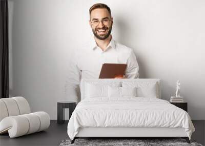 Confident business man holding digital tablet and smiling, standing against white background Wall mural