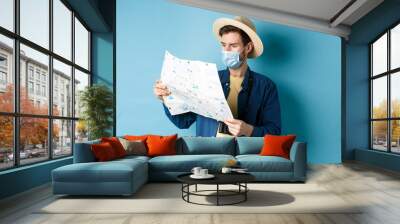 Covid-19, pandemic and travel concept. Handsome young guy in face mask and summer hat looking at tourist map, travelling on vacation, standing on blue background Wall mural