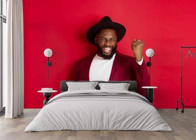 Fashion and party concept. Excited Black man rejoicing of winning, making fist pump gesture and say yes with satisfaction, standing against red background Wall mural