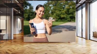 Fitness woman with water bottle and smartphone, jogging in park and smiling, looking at her mobile phone app, checking sport application Wall mural