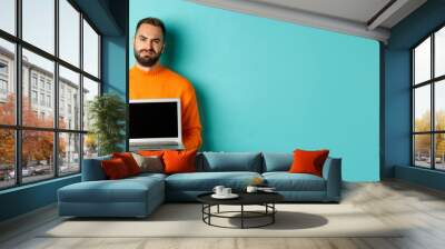 Handsome bearded man in orange sweater showing laptop screen, demonstrating promo, grimacing disappointed and upset standing over light blue background Wall mural