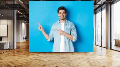 Handsome caucasian man in casual outfit, pointing fingers left and smiling, showing promo offer, standing over blue background Wall mural