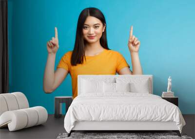 Heads up see awesome promo. Assertive good-looking confident asian woman showing friend advertisement, raise index fingers pointing top, smiling camera self-assured, stand blue background determined Wall mural