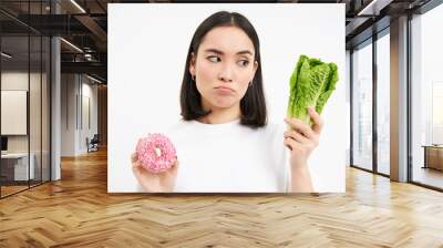 Healthy lifestyle concept. Young woman deciding between healthy vegetables, cabbage and delicious glazed doughnuts, white background Wall mural