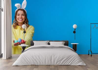 Holidays, people and fun concept. Charismatic cute blond girl holding coloured Easter eggs, wearing bunny ears and smiling joyfully, congratulate family with holy day, blue background Wall mural