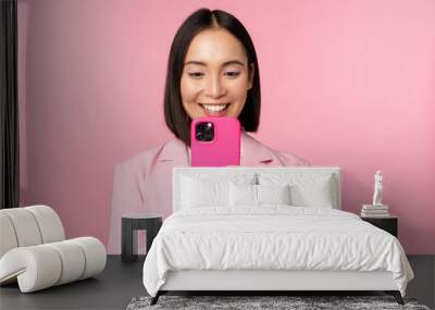 Image of smiling asian corporate woman in suit looking, watching on smartphone app, using mobile phone application, standing over pink background Wall mural