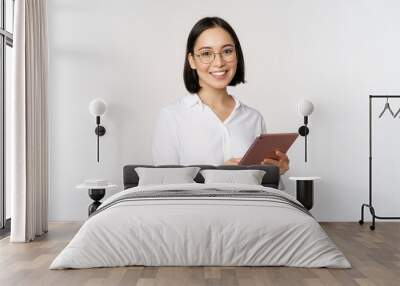 Image of young asian woman, company worker in glasses, smiling and holding digital tablet, standing over white background Wall mural