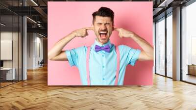 Irritated young man block noise, shut ears with hands and screaming annoyed, being disturbed by loud sound, standing over pink background Wall mural