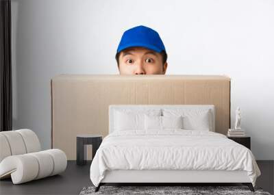 Online shopping, fast shipping concept. Surprised cute asian male courier, delivery man in blue cap, peeking behind large box with client order, bring package to customer doorstep, white background Wall mural