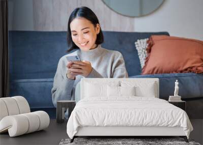 People and technology. Smiling asian woman sitting at home, using her mobile phone, typing message, browing internet or shopping online from smartphone app Wall mural